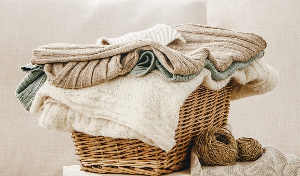 A wicker basket filled with folded knit sweaters in neutral colors, with two balls of yarn placed beside the basket.